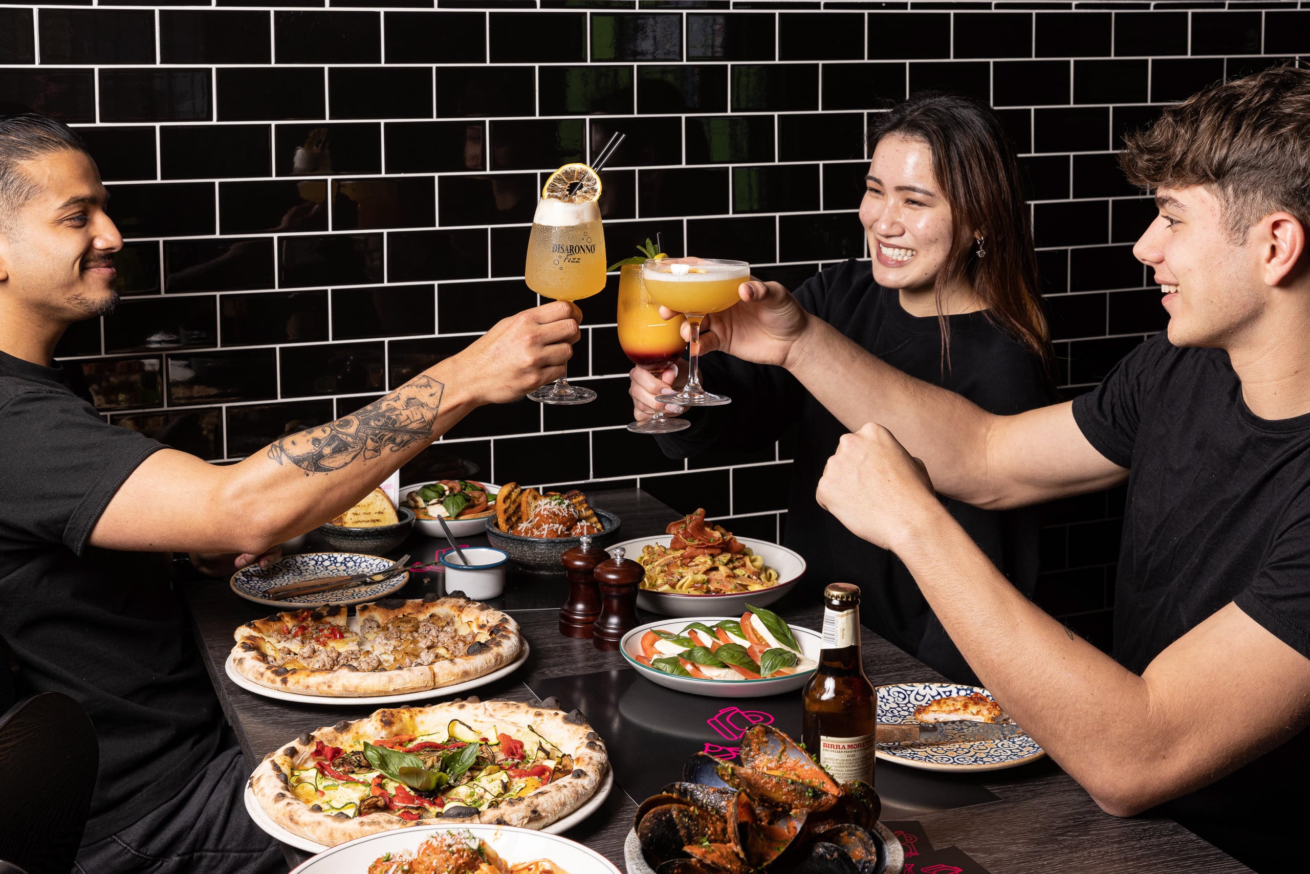 Happy people sit around a restaurant table and clink their cocktail glasses. The table is covered with pizza and pasta dishes.